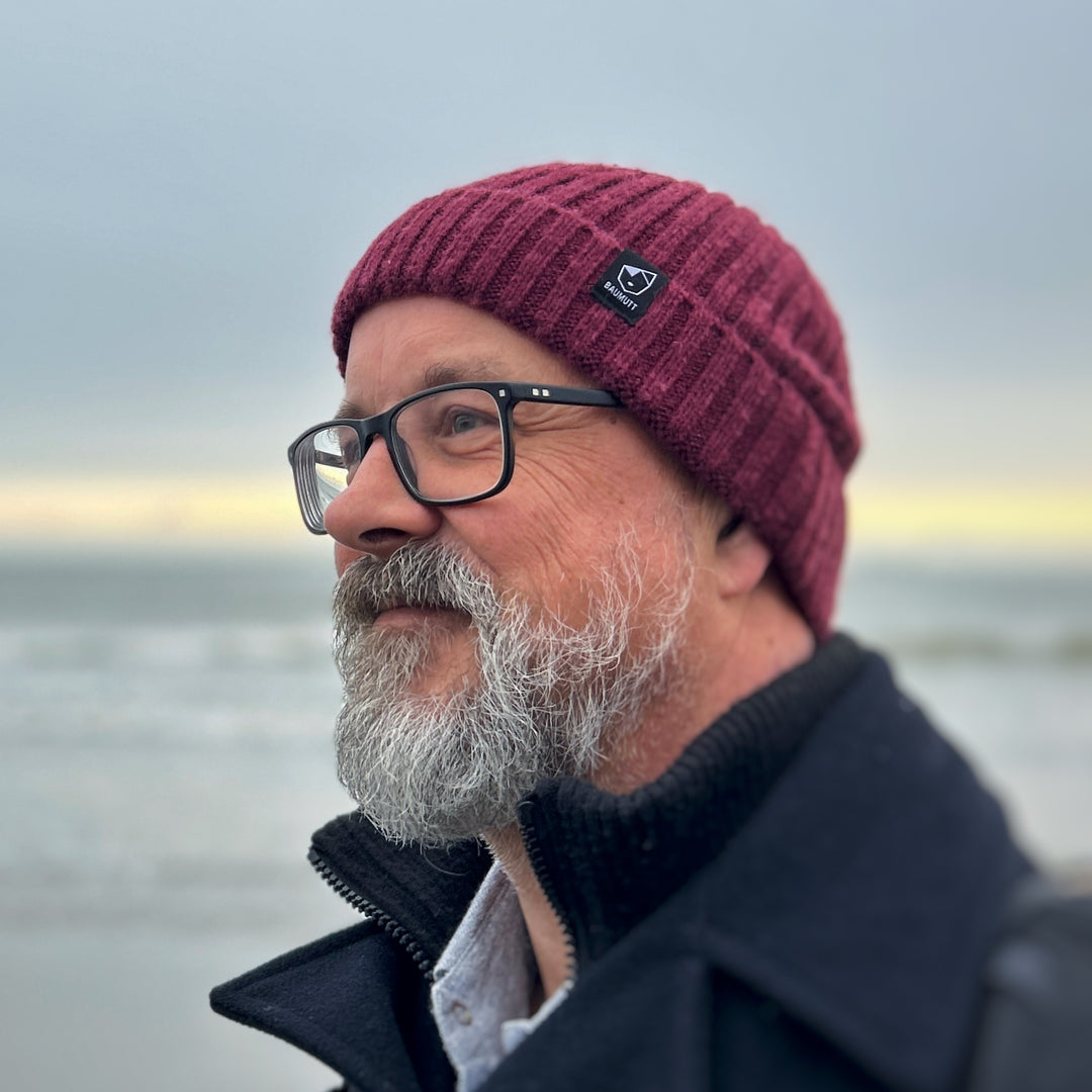 A burgundy red BAUMUTT fisherman beanie hat with a snug fit, designed for men and women. side view on male model