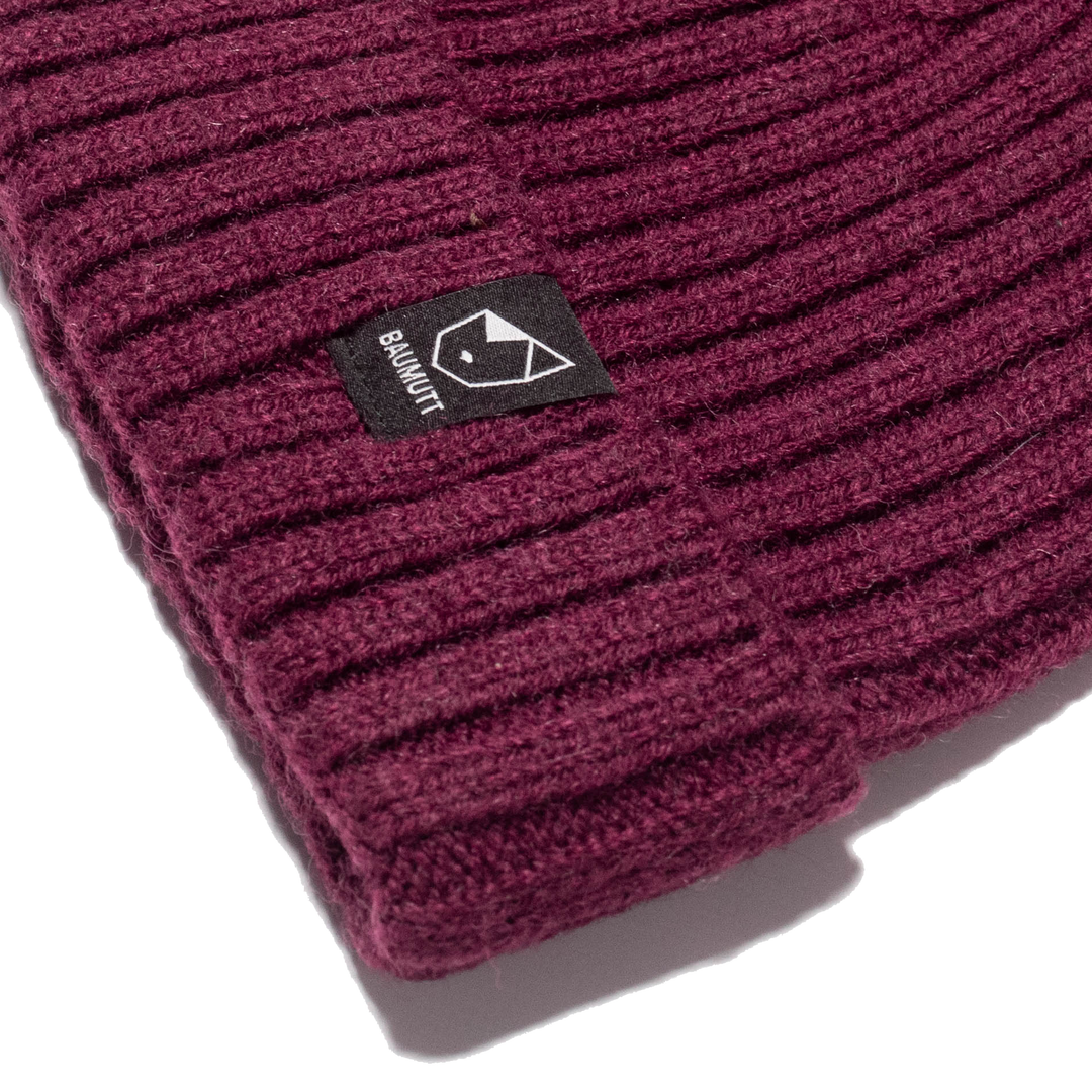 A Burgundy red BAUMUTT fisherman beanie hat with a snug fit, designed for men and women. side corner logo view
